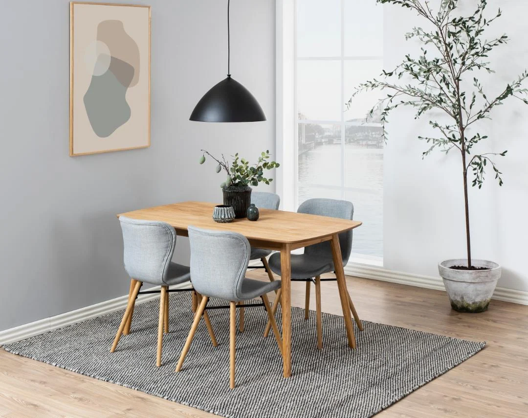 Een moderne eethoek met de Eettafel Nieuwe Sfeer 150 x 80 CM Rond in eikenhout naturel, vier grijze stoelen, een zwarte hanglamp erboven, plant in pot, groot raam met uitzicht op water, abstracte muurkunst en grijs vloerkleed op houten vloer.