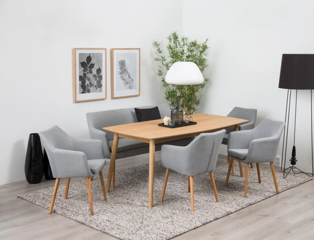 Een moderne eetkamer met de Eettafel Nieuwe Sfeer 150 x 80 CM Rond in naturel eiken, vijf grijs gestoffeerde stoelen, een witte lamp, een plant in pot, botanische prints op een witte muur en een grote zwarte vloerlamp op een vloerkleed met structuur.