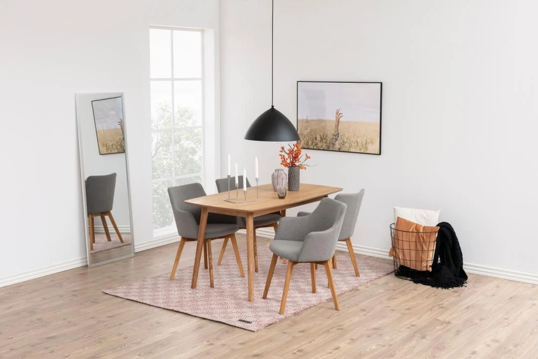 Een moderne eetkamer met de Eettafel Nieuwe Sfeer 150 x 80 CM Rond in naturel eiken, vier grijze stoelen, een hanglamp, een grote spiegel, wandkunst en een draadmand met kussens en een deken op een vloerkleed met patroon op hardhouten vloeren.