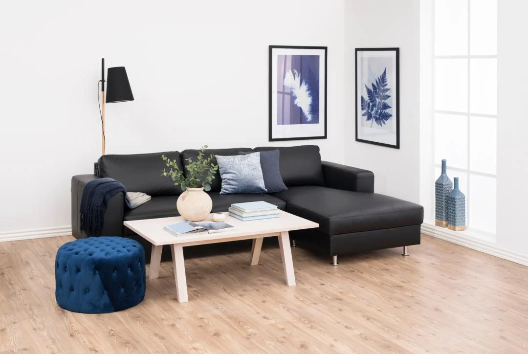 Een moderne woonkamer met een zwarte sofa, blauwe voetenbank, de Salontafel Roest in wit (70 x 45 cm) met rechthoekig eikenfineer, versierd met boeken en een vaas, blauwe muurkunst, een staande lamp en grote ramen die natuurlijk licht binnenlaten.