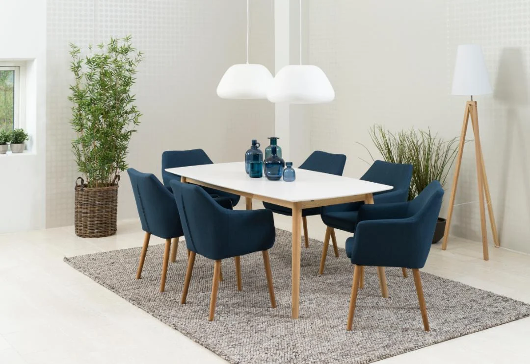 Een lichte, minimalistische eetkamer met de Eettafel Nieuwe Sfeer 150 x 80 CM rechthoek eikenhout tafel, zes blauw gestoffeerde stoelen, blauwe glazen vazen, hangende witte lampen, een grijs vloerkleed en groene planten.