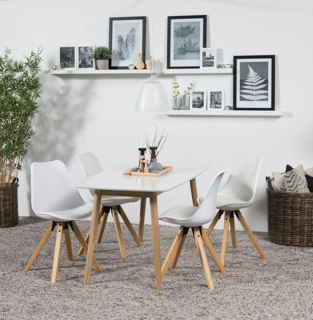 Een moderne eethoek is voorzien van de Eettafel Nieuwe Sfeer 150 x 80 CM, een rechthoekige eiken tafel, vier witte stoelen met houten poten, een grijs vloerkleed, planten, een mand met kussens en botanische kunst op witte muren en zwevende planken.