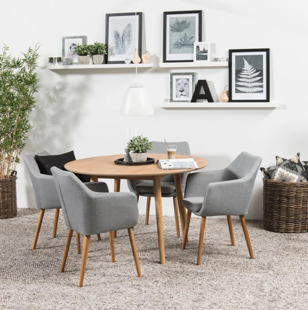 De Eettafel Nagano 120 CM Rond Eikenhout wordt gecombineerd met vier grijs gestoffeerde stoelen op een geweven tapijt, omringd door planten, manden en planken met ingelijste kunst en decor, waardoor een moderne, minimalistische eetruimte ontstaat.