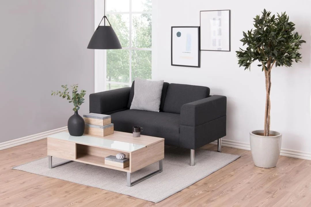 Een moderne woonkamer met een donkergrijze bank, grijs kussen en een Salontafel Wit 110 x 60 CM Rechthoek van kunststof op een vloerkleed, plus een boom in pot, muurkunst, een hanglamp en een groot raam met groen buiten.