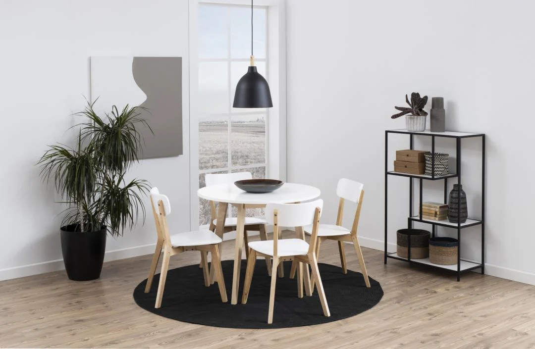 Een moderne eethoek is voorzien van de Eettafel Nieuwe Sfeer 90 CM Rond MDF Massief, vier houten stoelen, een zwarte hanglamp, een potplant, muurkunst, een raam en een zwarte metalen plank met decor op een lichthouten vloer.