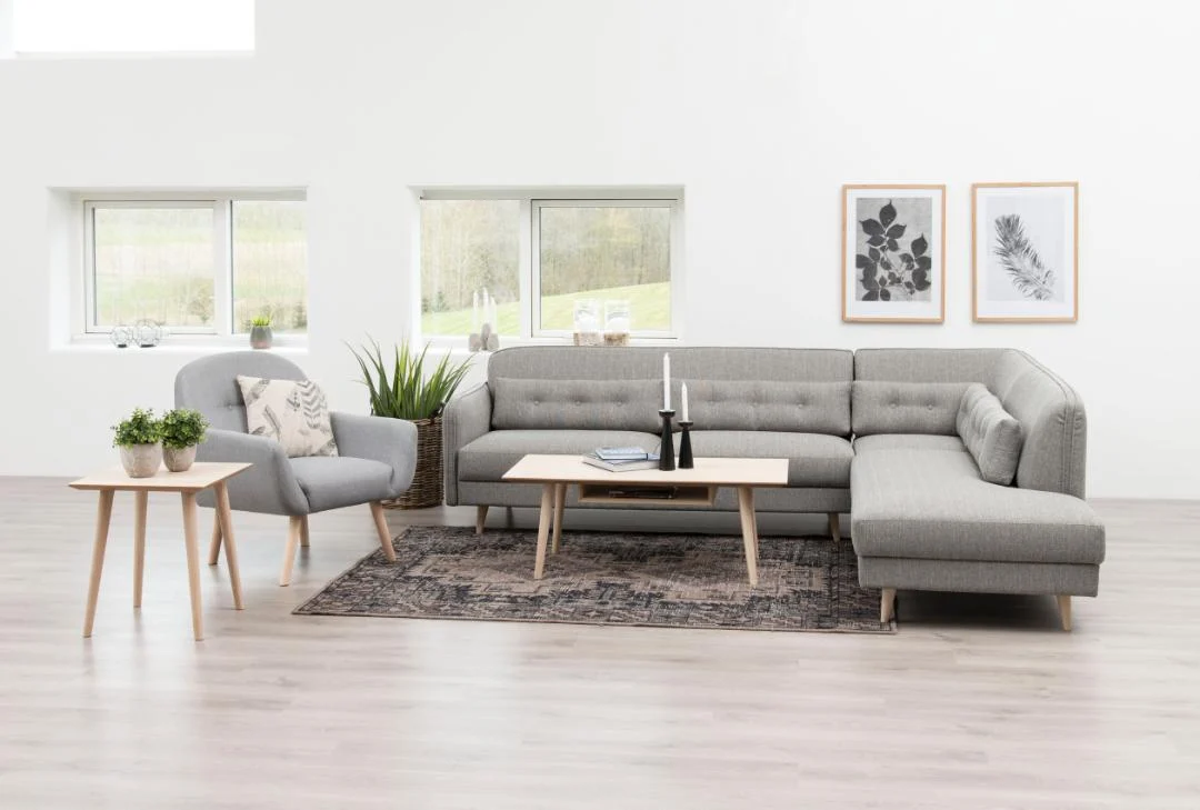 Een moderne woonkamer met een grijze sectionele bank, bijpassende fauteuil, Salontafel Wit 60 x 42 CM Rond in eikenfineer, planten en ingelijste botanische prints op een witte muur. Grote ramen brengen natuurlijk licht binnen en een vloerkleed met patroon bedekt de vloer.