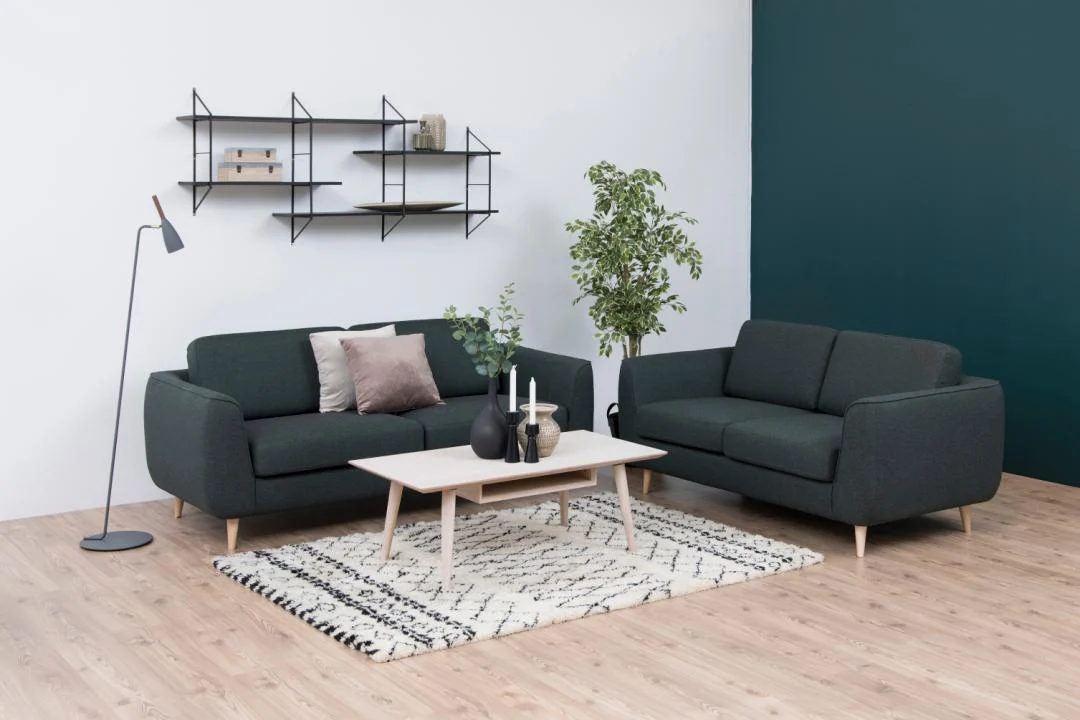 Moderne woonkamer met twee donkergroene banken, een ronde witte salontafel (Salontafel Wit 60 x 42 CM Rond) met eikenfineer, decoratieve items, een vloerkleed met patroon, staande lamp, wandplanken, een plant en een tweekleurige witte en donkergroene muur.