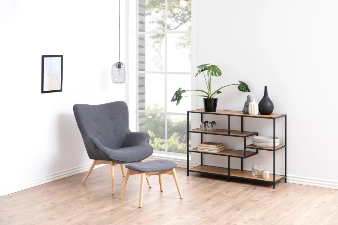 Een moderne woonkamer met een grijze fauteuil, bijpassende voetenbank, de Seaford boekenkast (een zwart metalen en houten plank) met planten en decor, een hanglamp en een groot raam met uitzicht op de buitenlucht.