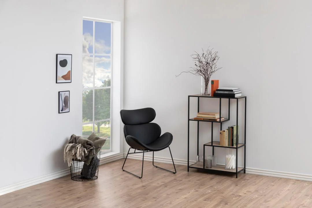Een moderne, minimale leefruimte heeft een zwarte stoel, de Seaford boekenkast met boeken en decor, een mand met dekens, twee kleine ingelijste kunstwerken aan de muur en een hoog raam dat natuurlijk licht binnenlaat.