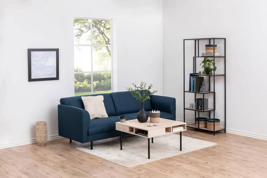 Een moderne woonkamer heeft een blauwe bank, een lichthouten salontafel, een wit vloerkleed, een Seaford boekenkast met decor, ingelijste kunst, een raam met groen buiten, lichte muren en een houten vloer.