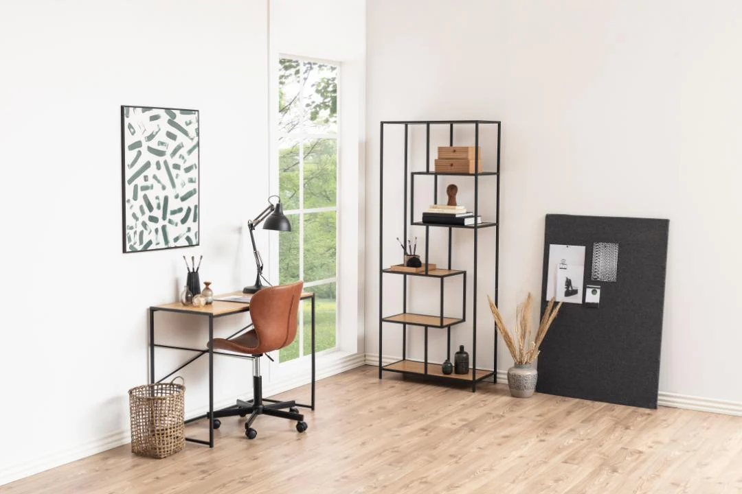 Minimalistisch kantoor aan huis met een klein bureau, bruine stoel, lamp, mand, muurkunst, prikbord en decor dat is uitgestald op de Seaford boekenkast van zwart metaal, tegen witte muren met een groot raam met uitzicht op groen.