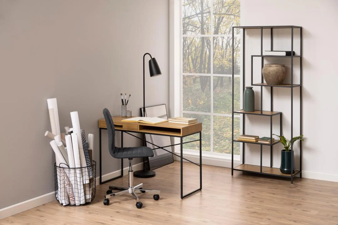 Een modern minimalistisch kantoor aan huis met een bureau, stoel, lamp, boeken en decor. Natuurlijk licht stroomt naar binnen door een groot raam. In een mand liggen opgerolde papieren en in de Seaford boekenkast staan decoratieve spullen. Houten vloer en neutrale muren.