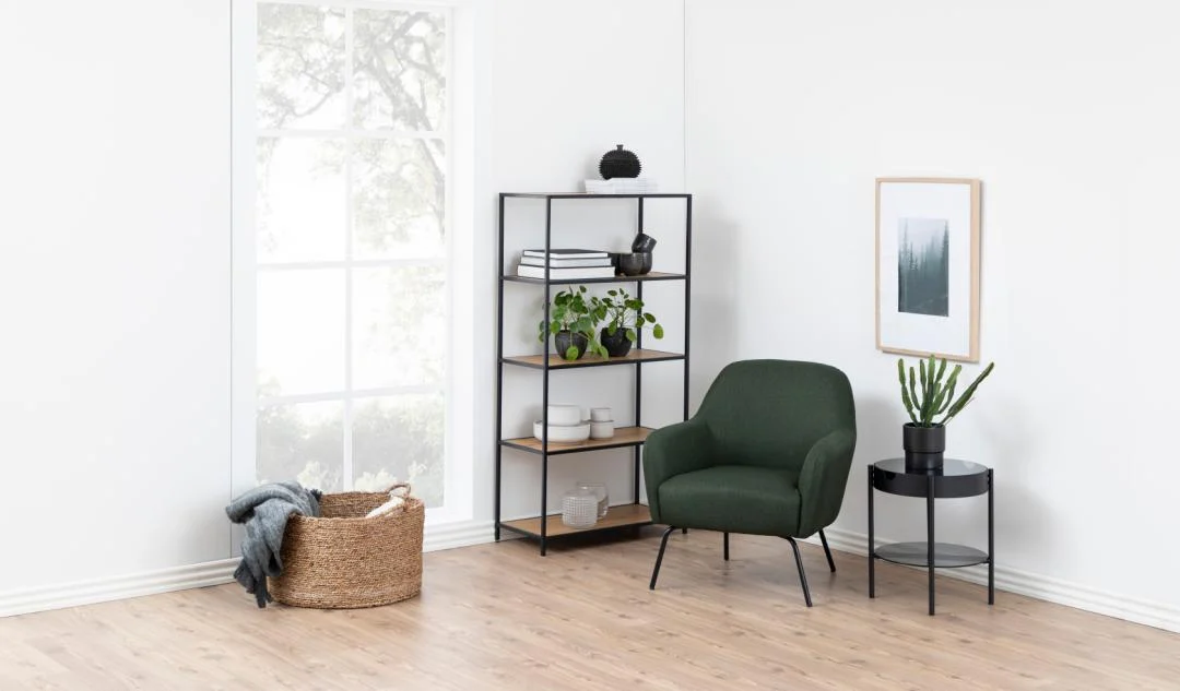 Een moderne minimalistische woonkamerhoek met een groene fauteuil, zwarte ronde bijzettafel met een potplant, Seaford boekenkast met decor, ingelijste kunst aan de muur, groot raam en een gevlochten mand op de houten vloer.