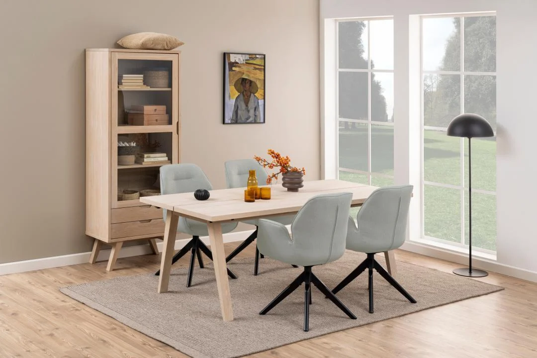 Een moderne eetkamer is voorzien van de Eettafel Nieuwe Sfeer 200 x 95 CM rechthoek spaanplaat tafel, vier grijze stoelen, een beige vloerkleed, een houten kast met boeken en decor, een staande lamp, een raam met groen en een schilderij boven de kast.