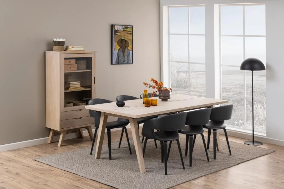 Een moderne eetkamer met de Eettafel Nieuwe Sfeer 200 x 95 CM rechthoekige spaanplaattafel, zes zwarte stoelen, een beige vloerkleed, een houten kast met boeken en servies, een staande lamp, muurkunst en grote ramen met een besneeuwd uitzicht.