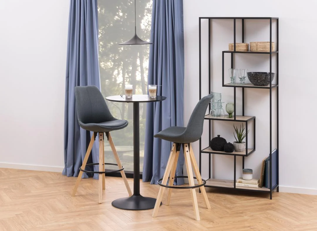 Een moderne eethoek met twee grijze barkrukken aan een ronde zwarte tafel bij een raam met blauwe gordijnen, drankjes bovenop. De Seaford boekenkast toont glaswerk en decor tegen de witte muur.