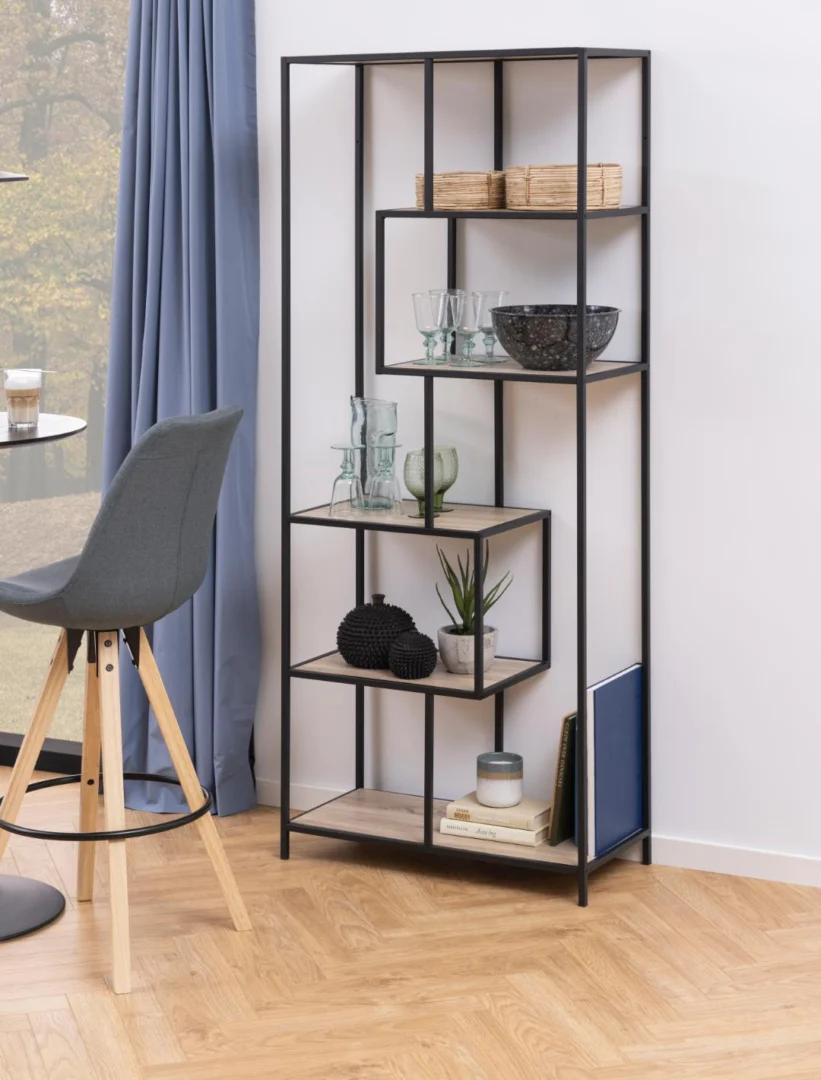 De Seaford boekenkast, een zwart metalen rek met lichthouten planken, toont glaswerk, schalen, manden, decor, een plant en boeken, en staat naast een grijze stoel in een moderne kamer met houten vloeren.