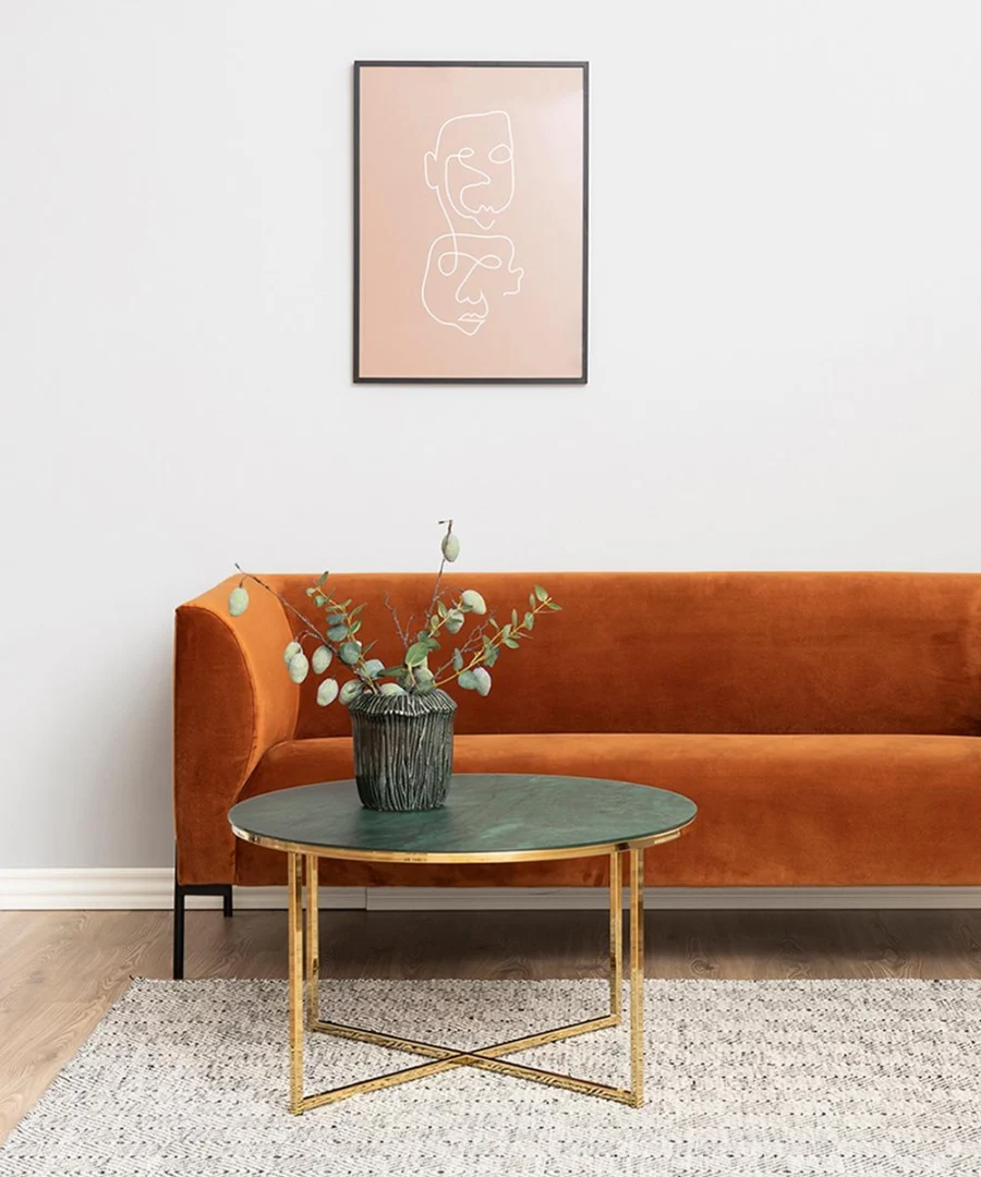 Een moderne woonkamer met een oranje fluwelen bank, de Salontafel Groen 80 CM Rond Kunststof salontafel met een potplant, een licht vloerkleed met structuur en een minimalistisch lijnkunstportret aan de muur.