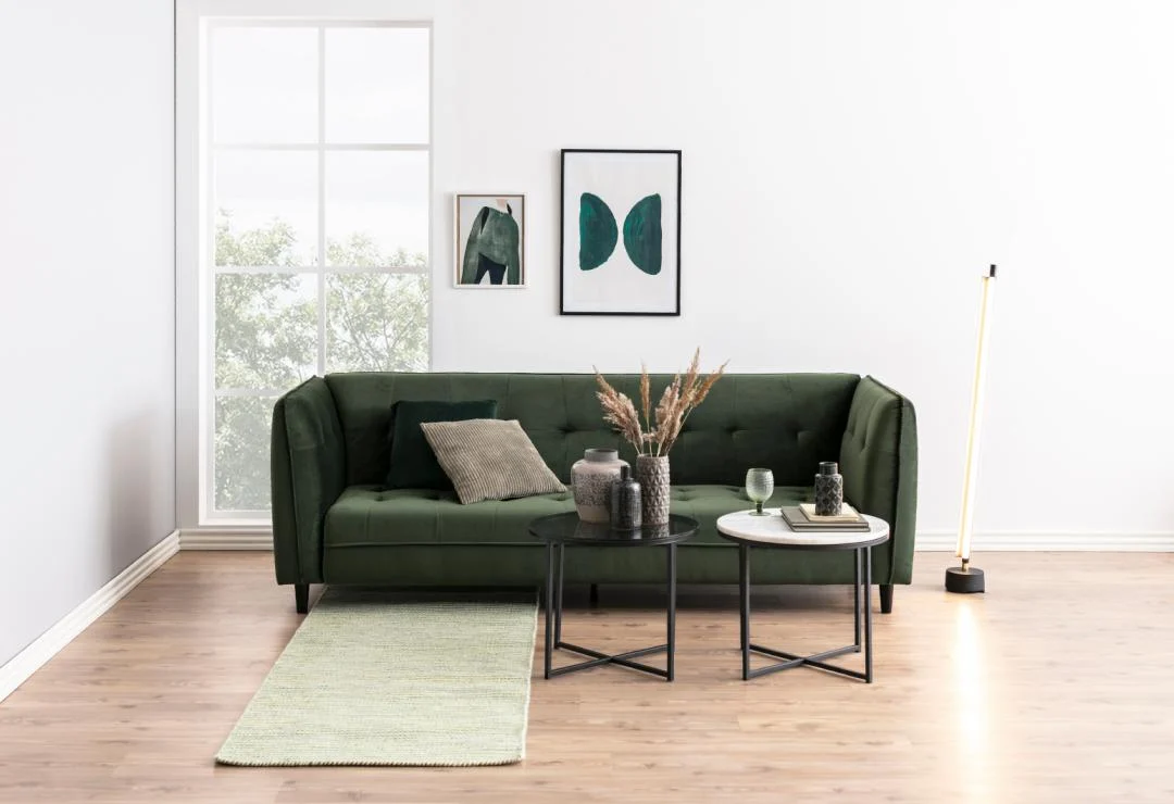 Een moderne woonkamer met een donkergroene bank, een Bijzettafel Grijs 55 CM Rond Kunststof salontafel met decor, een staande lamp, een groen vloerkleed en ingelijste kunstwerken op witte muren, bij een groot raam met uitzicht op bomen.