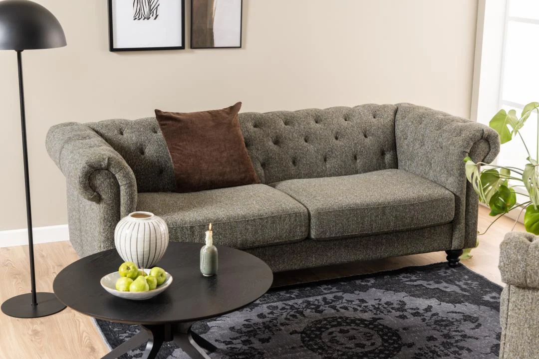 Een grijs getufte bank met een bruin kussen ligt op een vloerkleed met donker patroon in een moderne woonkamer. In de Salontafel Zwart 80 CM Rond Eikenfineer staan een gestreepte vaas, een kleine fles en groene appels. Vlakbij staat een zwarte vloerlamp.