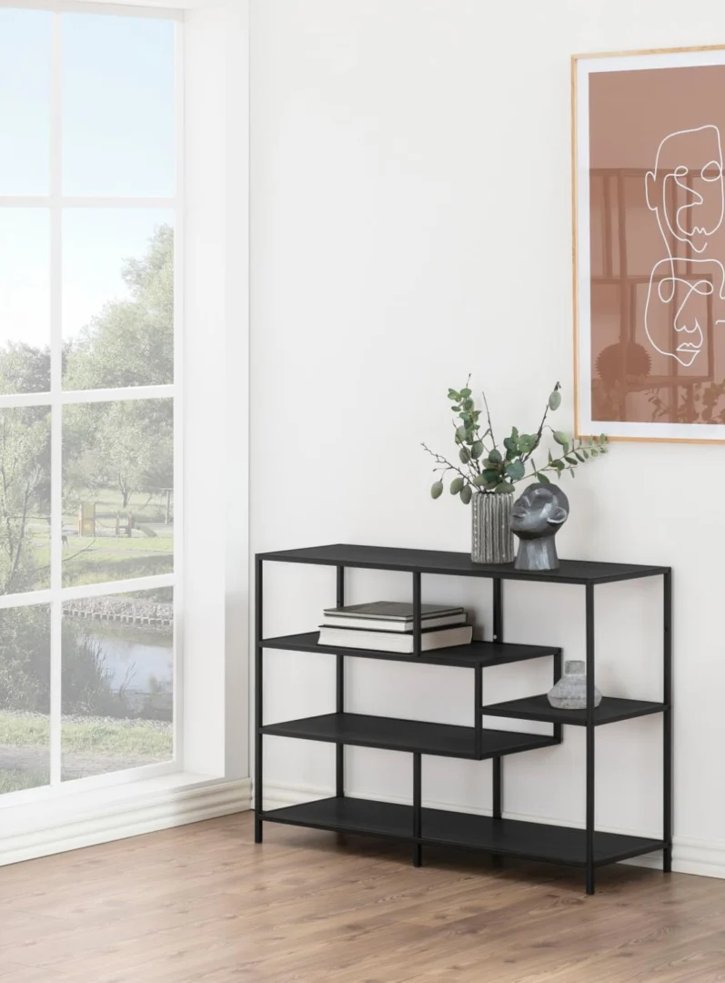 De Seaford boekenkast, een moderne zwarte metalen plank, toont boeken, groen in een vaas en een grijze decoratieve kop tegen een witte muur bij een groot raam en abstracte kunst in een lichte kamer met houten vloer.