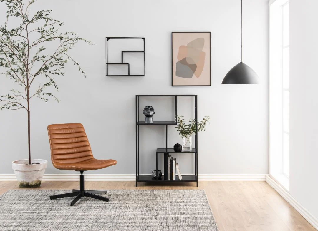 Moderne minimalistische woonruimte met een bruine leren stoel, Seaford boekenkast met decor, ingelijste abstracte kunst op witte muren, een boom in pot, geometrische wandplank en zwarte hanglamp boven een lichthouten vloer en grijs vloerkleed.