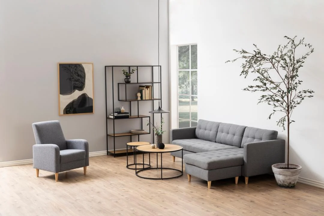 Een moderne woonkamer met een grijze fauteuil, sofa, ronde houten salontafels met zwarte metalen frames, een hoge potplant, Seaford boekenkast, abstracte muurkunst en een groot raam dat natuurlijk licht binnenlaat.