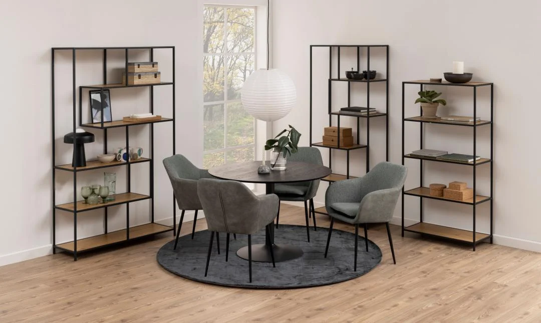 Een moderne eethoek met een ronde zwarte tafel, vier grijs gestoffeerde stoelen op een donker cirkelvormig vloerkleed, een houten vloer, een hanglamp met papieren lantaarn en twee Seaford boekenkasten met decor. Groot raam op de achtergrond.