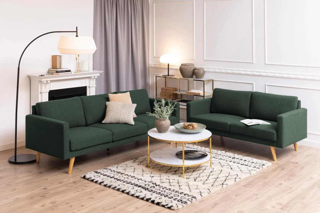 Een moderne woonkamer met twee donkergroene banken, een Salontafel Goud, Wit 80 CM Rond Kunststof met een plant, een vloerkleed met patroon, een staande lamp en planken met decor tegen witte wanden met panelen en een grijs gordijn.