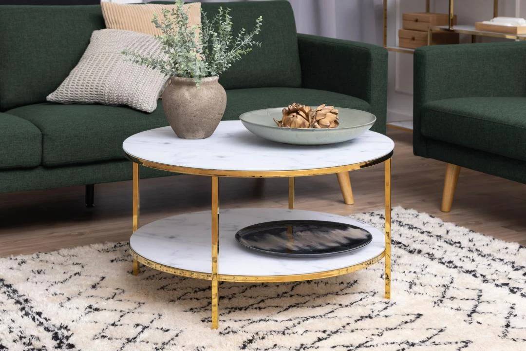 De Salontafel Goud, Wit 80 CM Rond Kunststof staat op een wit vloerkleed met een beige vaas met groen en een schaal met dennenappels. Groene banken en een neutrale inrichting maken de look op de achtergrond compleet.