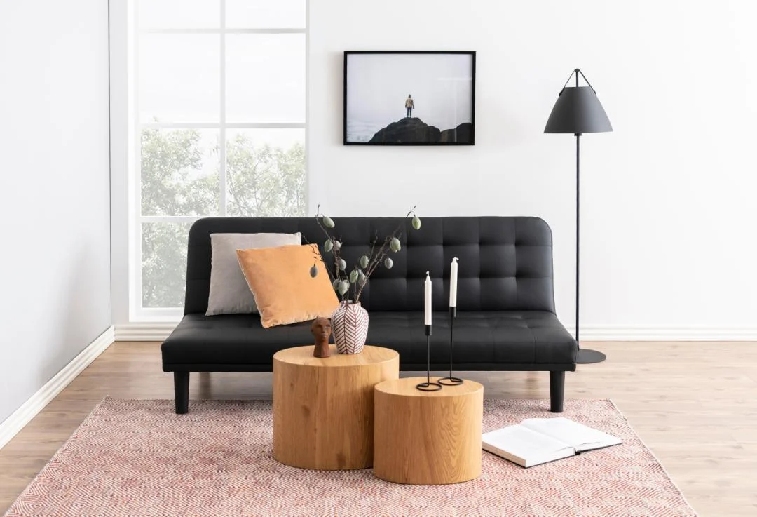 Een moderne woonkamer met een zwarte bank, beige en oranje kussens, twee salontafels Ovaal Kunststof 33 x 40 CM, een staande lamp, een vaas met takken, twee kaarsen, een open boek, een roze vloerkleed en een ingelijste foto aan de muur.