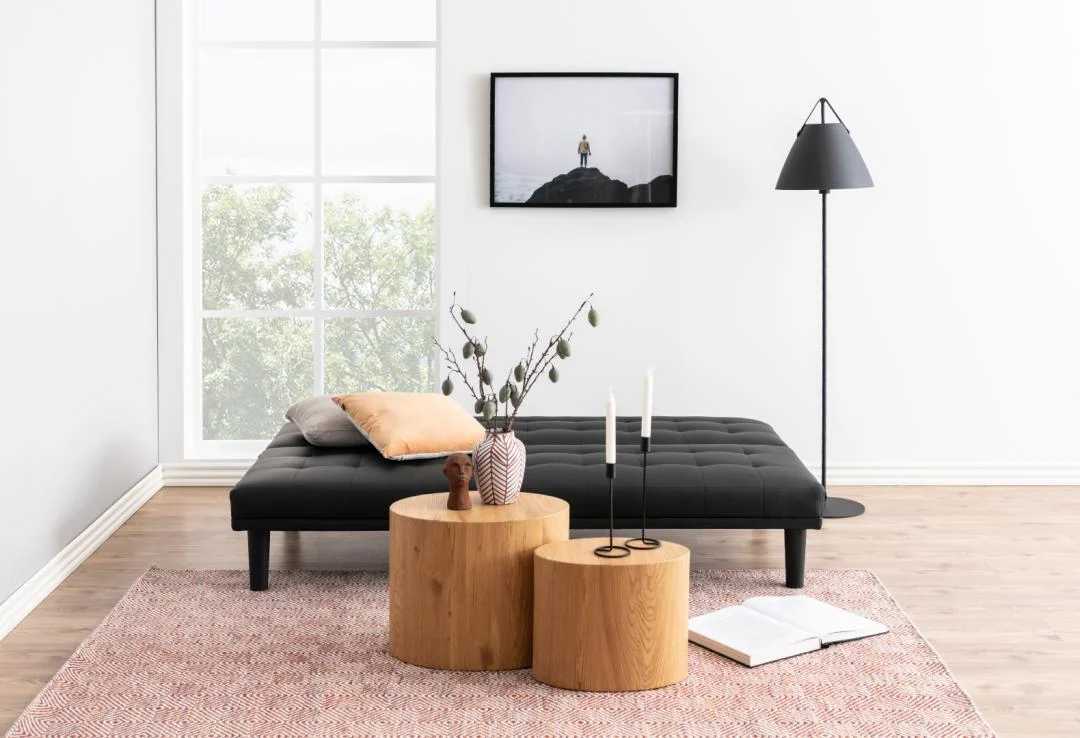 Minimalistische woonkamer met een zwarte futon, twee salontafels 33 x 40 CM van Ovaal Kunststof, decoratieve vazen, kaarsen, een open boek, een staande lamp, een ingelijste foto aan de muur en een groot raam dat natuurlijk licht binnenlaat.
