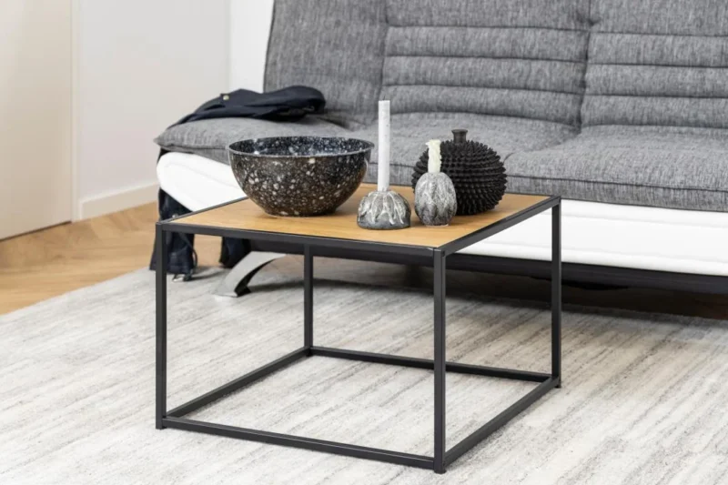 De Salontafel Zwart 60 x 60 CM Vierkant, gemaakt van compact kunststof, staat op een licht vloerkleed voor een grijze bank en toont een grote schaal, kaarsen en een bol met structuur als decoratieve accenten.