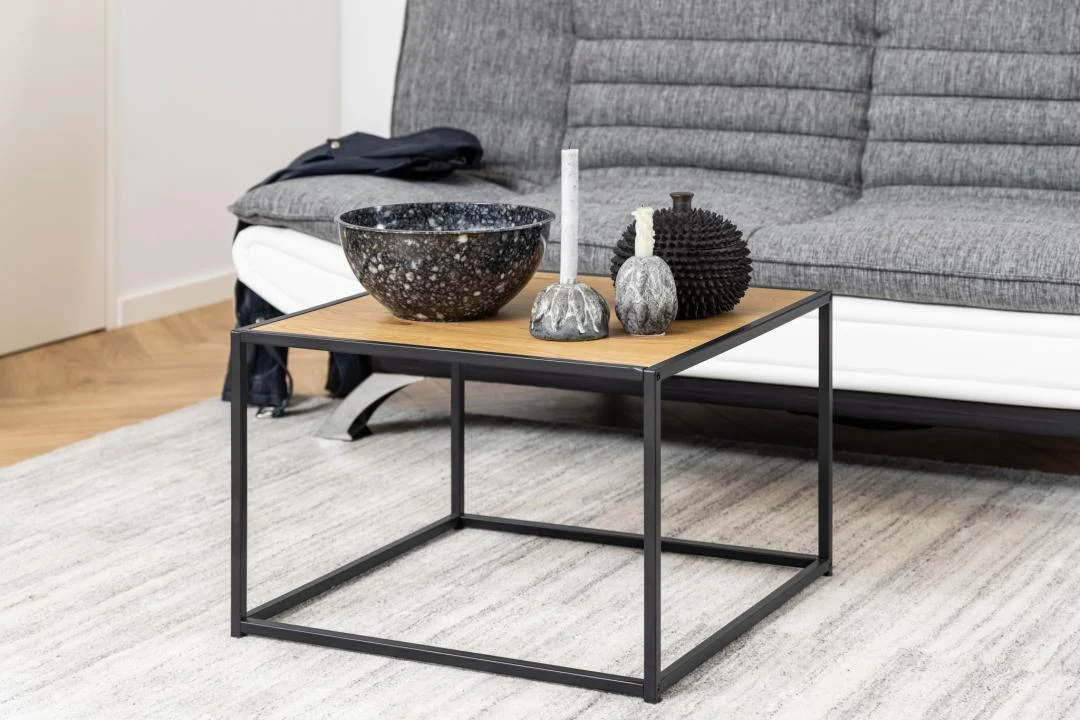 De Salontafel Zwart 60 x 60 CM Vierkant, gemaakt van compact kunststof, staat op een licht vloerkleed voor een grijze bank en toont een grote schaal, kaarsen en een bol met structuur als decoratieve accenten.