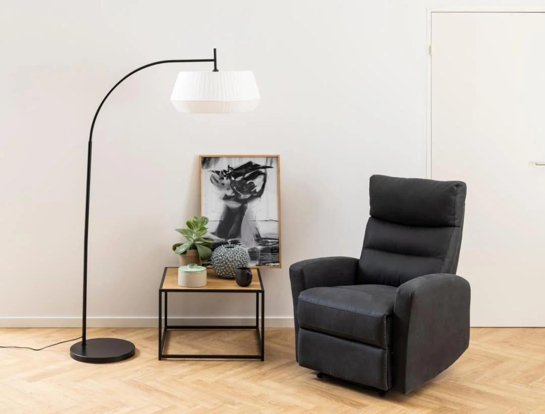 Een moderne woonkamerhoek met een zwarte fauteuil, een zwarte vloerlamp met witte kap en Salontafel Zwart 60 x 60 CM Vierkant (Kunststof Compact) als bijzettafeltje met planten en decor, plus abstracte kunst tegen een witte muur met een gesloten deur.