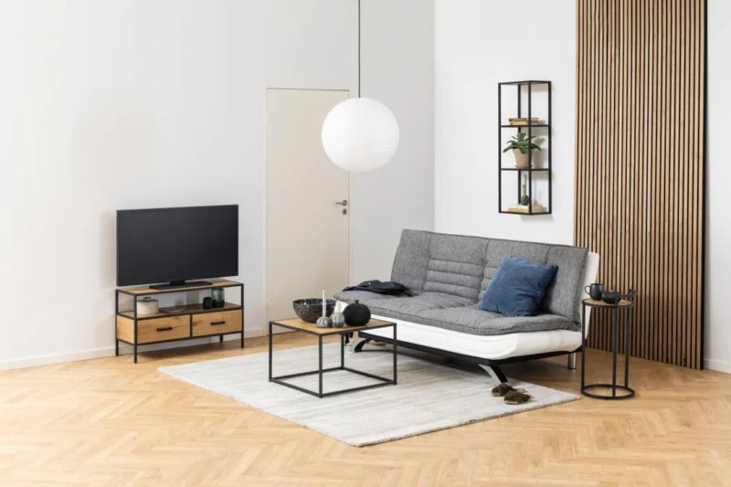 Een moderne woonkamer met een grijze futonbank, blauwe en grijze kussens, een tv op een zwarte en houten standaard, een Salontafel Zwart 60 x 60 CM Vierkant (kunststof compact), ronde witte hanglamp, lamellen wandaccenten en een licht vloerkleed op visgraat houten vloer.