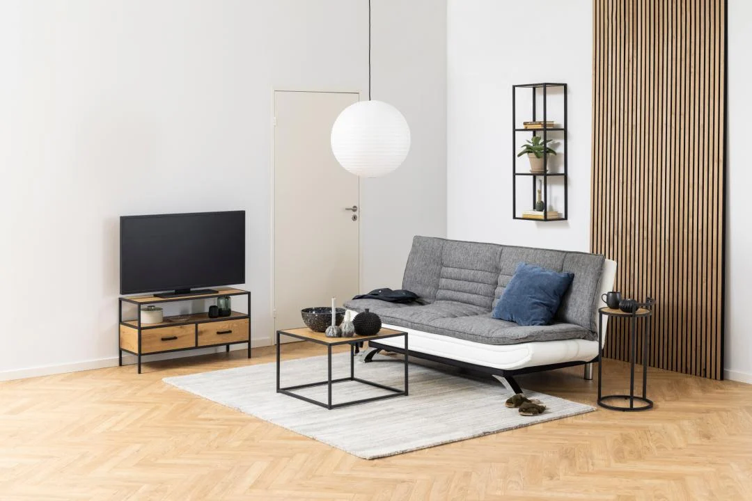 Een moderne woonkamer met een grijze futonbank, blauwe en grijze kussens, een tv op een zwarte en houten standaard, een Salontafel Zwart 60 x 60 CM Vierkant (kunststof compact), ronde witte hanglamp, lamellen wandaccenten en een licht vloerkleed op visgraat houten vloer.