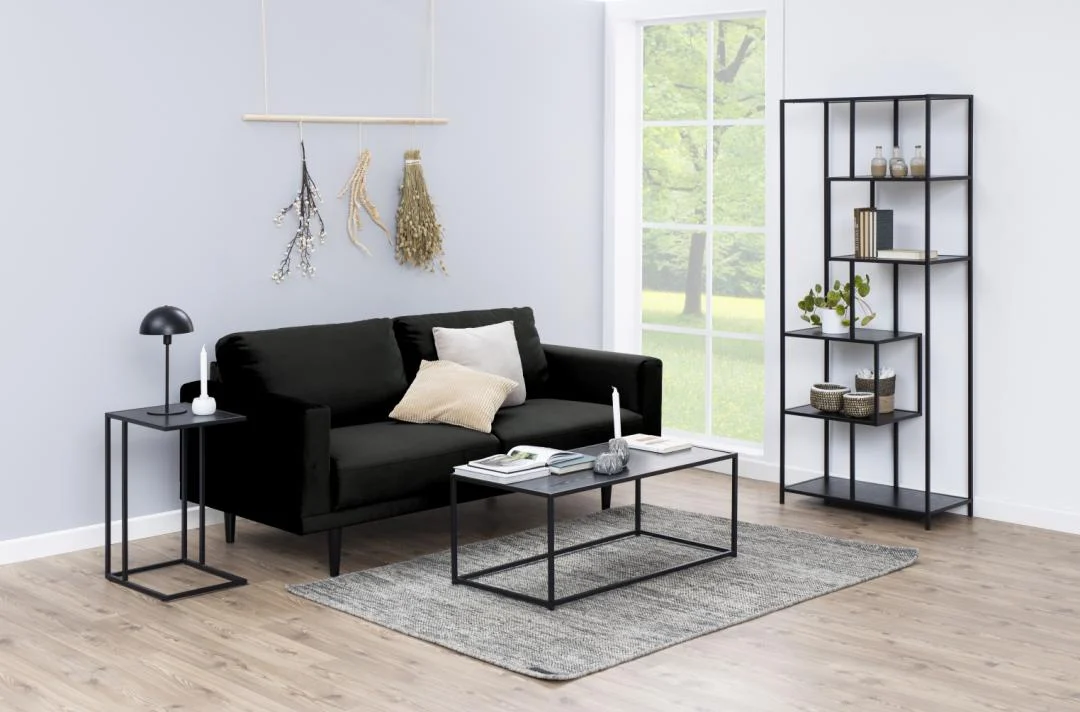 Een moderne woonkamer met een zwarte bank, lichte kussens, een glazen salontafel, een Bijzettafel 35 x 63 CM Rechthoek kunststof met een zwarte lamp, een grijs vloerkleed, minimalistische plank, decoratieve wandkleden en een groot raam met groen.