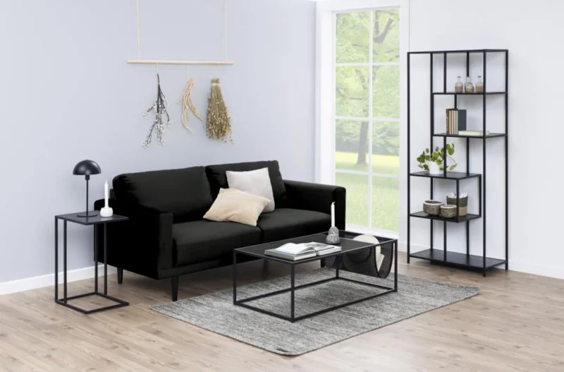 Een moderne woonkamer met een zwarte bank, lichte kussens, glazen salontafel, de Bijzettafel 35 x 63 CM Rechthoek kunststof bijzettafel met lamp, een hoge zwarte stellingkast met decor, grijs vloerkleed op lichthouten vloer en groot raam met groen.