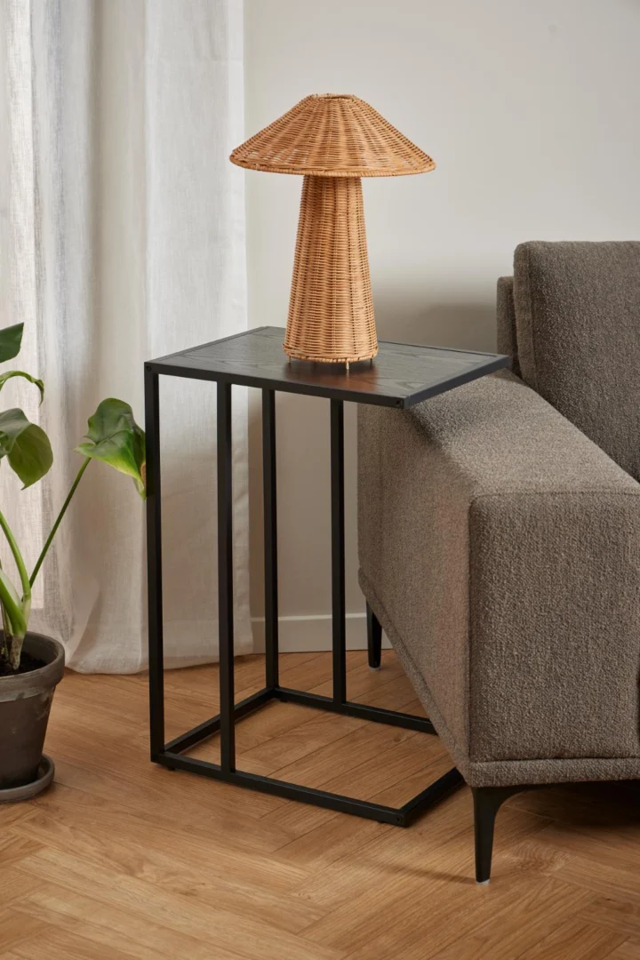 Een gevlochten rotan tafellamp staat op de Bijzettafel 35 x 63 CM Rechthoek (kunststof) naast een bank met grijze structuur. Een potplant staat vlakbij en een wit gordijn hangt bij de lichte houten vloer.