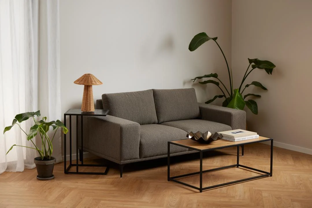 Een moderne woonkamer met een grijze bank, een zwart metalen salontafel met boeken en decor, de Bijzettafel 35 x 63 CM Rechthoek (kunststof) als bijzettafel, twee potplanten, lichthouten visgraatvloeren en witte gordijnen.
