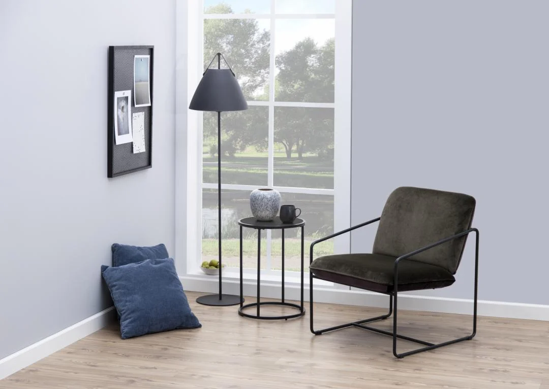 Een moderne minimalistische leeshoek met een zwarte fauteuil, een Bijzettafel Zwart 40 CM Rond Kunststof die een vaas en beker vasthoudt, een zwarte staande lamp, twee blauwe kussens op de vloer en een prikbord naast een groot raam met een schilderachtig uitzicht.