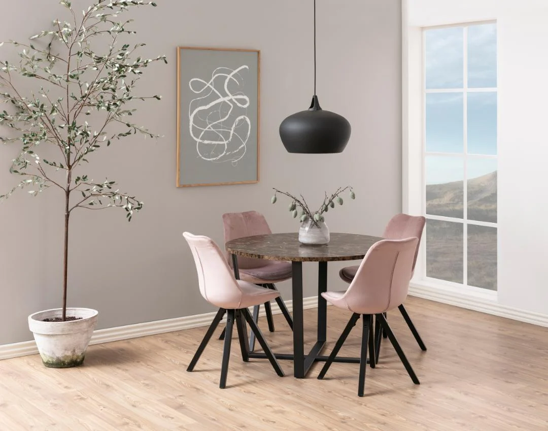 Een moderne eethoek met de Eettafel Nieuwe Sfeer 110 CM Rond MDF staal rond tafel, vier roze stoelen, een boom in pot, zwarte hanglamp, abstracte muurkunst en grote ramen met uitzicht op de bergen.