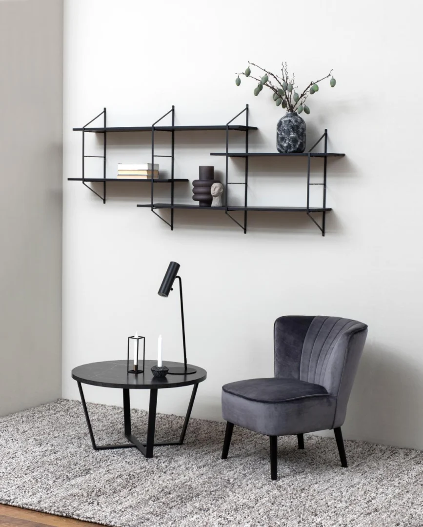 Een moderne leefruimte met een grijze fluwelen stoel, de Salontafel Zwart 77 CM Rond Kunststof met kandelaars en een lamp, een grijs vloerkleed met textuur en een geometrische zwarte wandplank met boeken, vazen en een decoratieve tak.