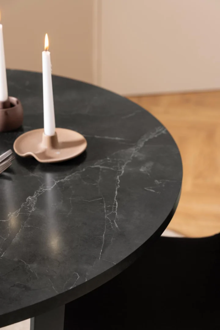 De Eettafel Amble Rond Marmer Metaal Mat met zwart onderstel houdt twee brandende witte kaarsen in beige houders. Een gedeeltelijke vork ligt op tafel en op de achtergrond verschijnt een houten vloer.