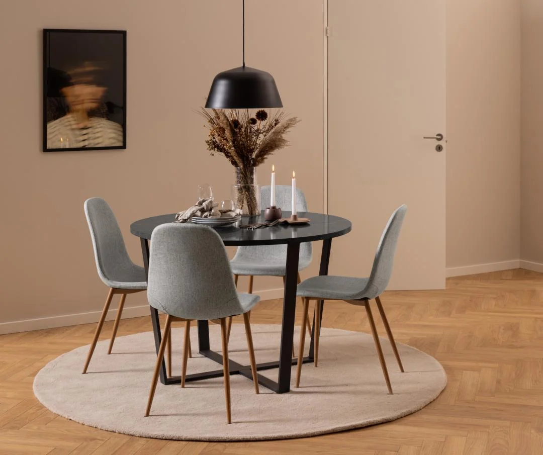 In een moderne eethoek staat de Eettafel Amble Rond Marmer Metaal met een zwart onderstel, omringd door vier grijze stoelen, droogbloemen in een vaas, brandende kaarsen en een zwarte hanglamp erboven, alles op een rond vloerkleed met een lichthouten vloer en beige muren.