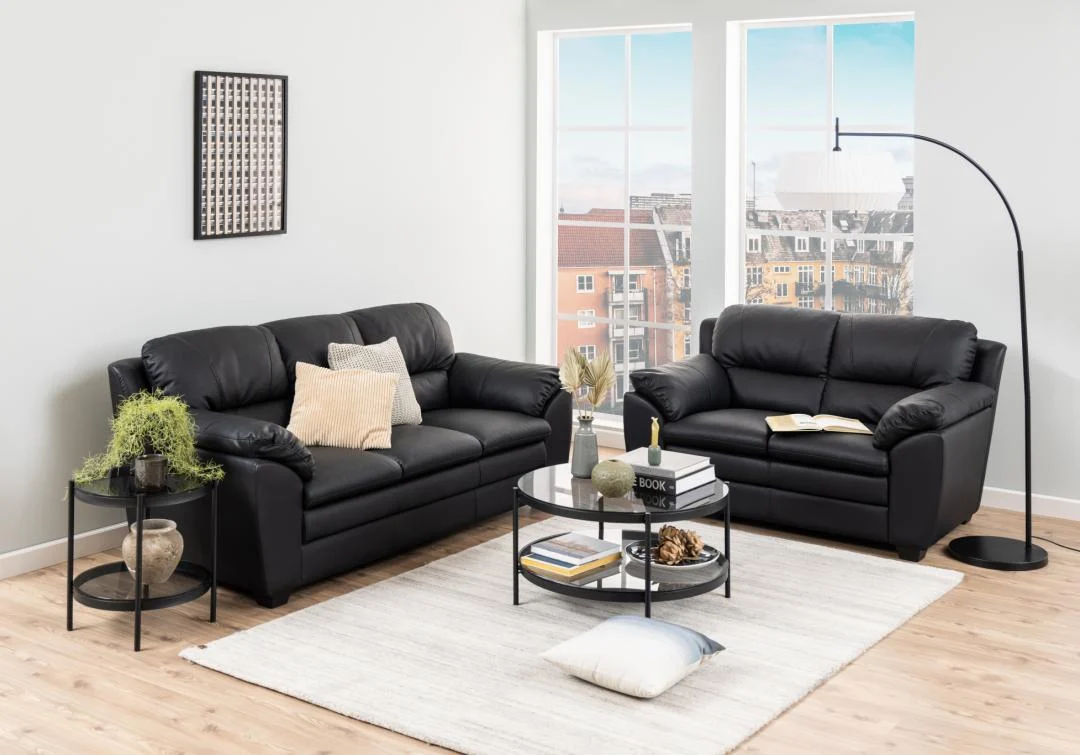 Een moderne woonkamer met twee zwarte leren banken, de Salontafel Zwart 79 CM Rond Kunststof als ronde salontafel, twee bijzettafels met planten, een staande lamp, grote ramen met een stadsgezicht en een wit vloerkleed op een lichthouten vloer.