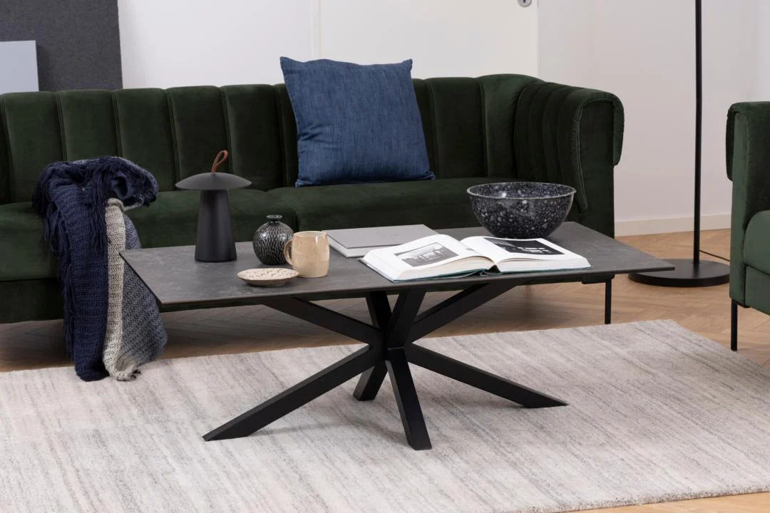 Een moderne woonkamer met een groene bank, blauw kussen en een Salontafel Zwart 130 x 70 CM Rechthoek Keramiek met een open boek, laptop, mok, kom, lamp en opgevouwen dekens op een licht vloerkleed op een houten vloer.