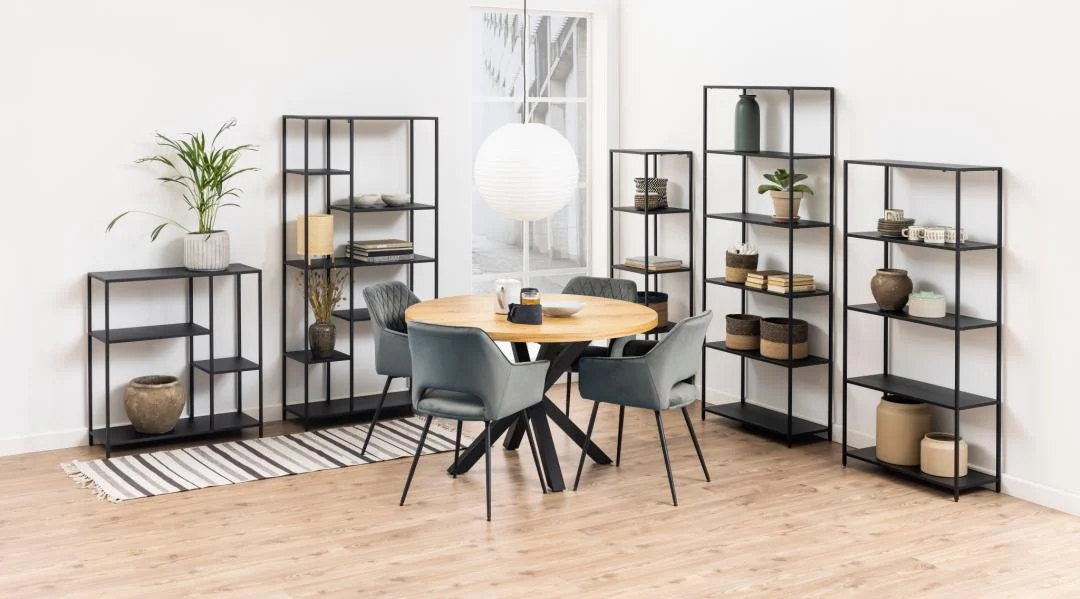 Lichte kamer met houten vloer en ronde tafel met grijze stoelen. Zwarte, metalen, asymmetrische boekenkast met zes planken.