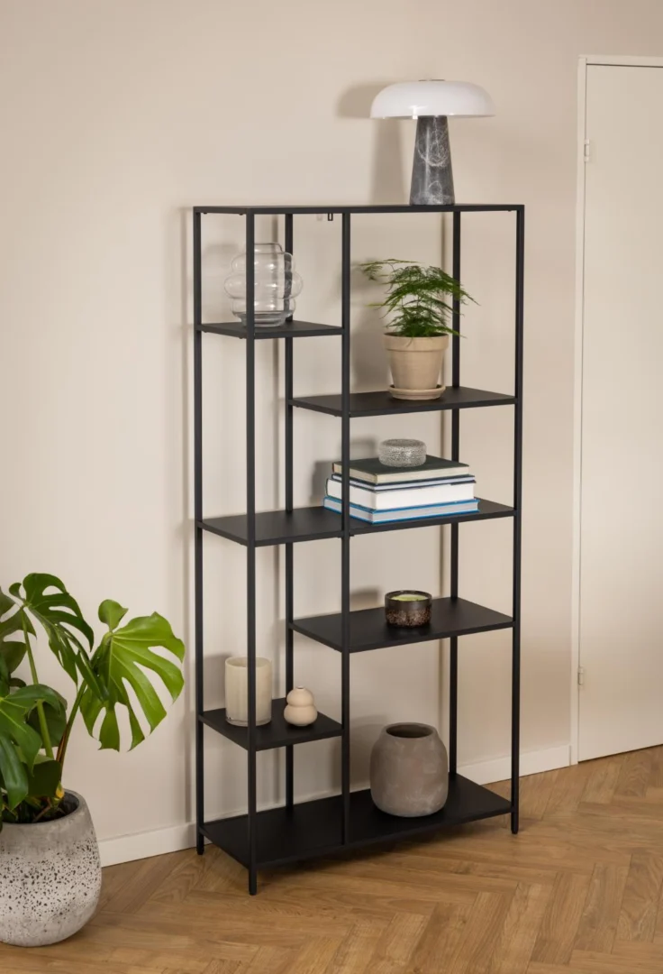 Zwarte, asymmetrische industriële boekenkast met zes planken tegen een crèmekleurige muur. Diverse decoraties en een kamerplant op de planken.